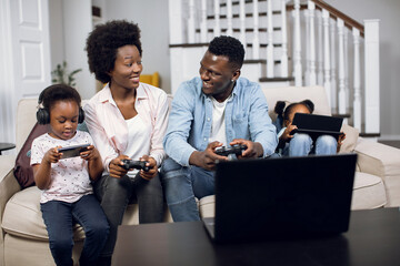 Two african sisters using tablet and smartphone while happy young parents playing games on laptop. Happy family with modern gadgets at home. Leisure and technology concept.