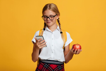 Happy young blonde girl pupil with pigtails and backpack in glasses typing on phone, holds red apple