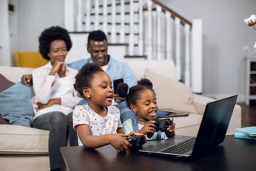 Two african sisters holding joysticks and playing games on modern laptop. Blur background of parents sitting on couch and using smartphone. Modern gadgets for lifestyles.