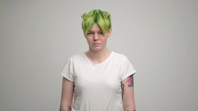 A young girl with a short pixie haircut and green hair in a white T-shirt on a light background. A woman poses in the studio, smiles and shows emotions. A breast portrait.
