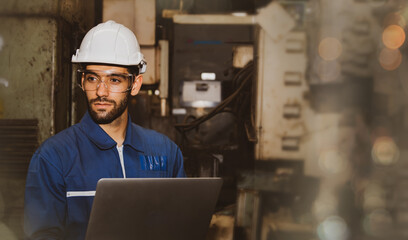 Professional handsome male engineer in blue uniform and helmet working on a laptop computer in the indoor field of a mechanical factory.