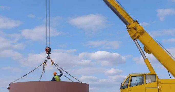 A team of workers in overalls and helmets help a large yellow truck crane move huge metal parts on a nearby truck.