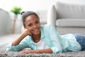 Smiling young pretty black girl student in casual looks at camera and rests, lies on floor in living room interior