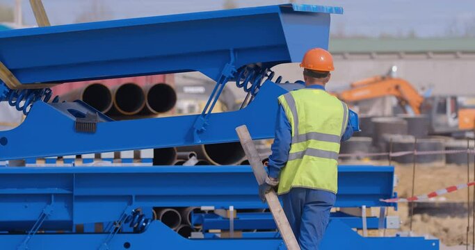 A Worker In A Yellow Vest And Orange Helmet Helps A Crane To Neatly Lay Down Large Blue Elements Of Factory Equipment