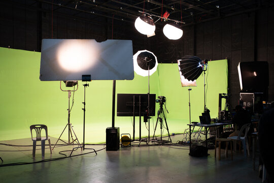 Behind the scenes of a green screen set with a video camera, monitor, and a set of many types of lighting equipments on tripods, set up for shooting video or broadcasting in a television studio.