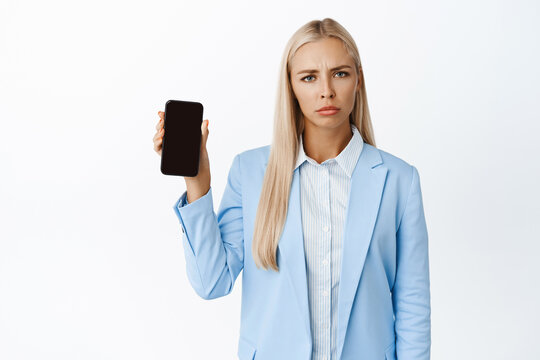 Image Of Displeased Corporate Woman, Frowning Upset And Showing Mobile Phone Screen, Standing In Blue Suit Over White Background