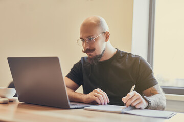 Young bald man working on laptop sitting at desk studying doing something notes. Distance Learning 