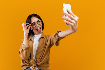 Playful woman taking selfie, showing tongue at cellphone camera and touching glasses ,posing over yellow background