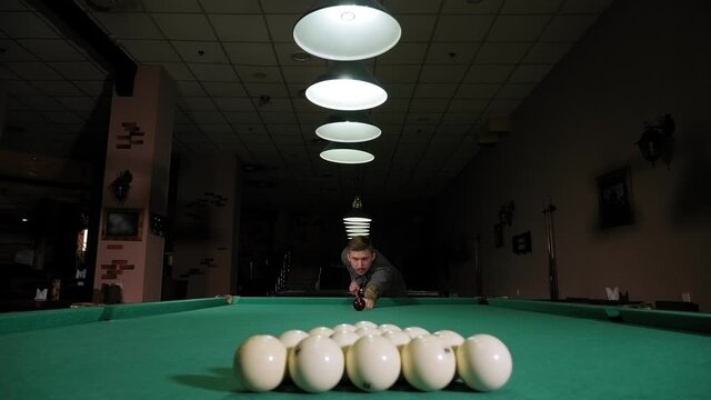 A young man playing Billiards in a club in the evening, he takes aim and prepares to hit the balls with a cue. A young man lines up his shot as he breaks the balls for the start of a game of billiards