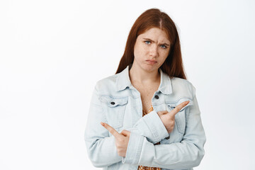 Indecisive redhead girl pointing sideways, choosing between two variants, troubled to make decision, standing over white background