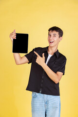 Portrait of young boy, teen or student in casual clothes, black shirt and blue jeans using tablet isolated on yellow studio backgroud.