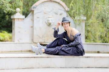 Casually dressed woman in a hat sits on the curb of the park decorative fountain made in the classical style