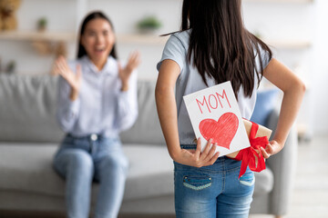 Mother's day gift. Girl congratulating excited mother with holiday, hiding greeting card and...