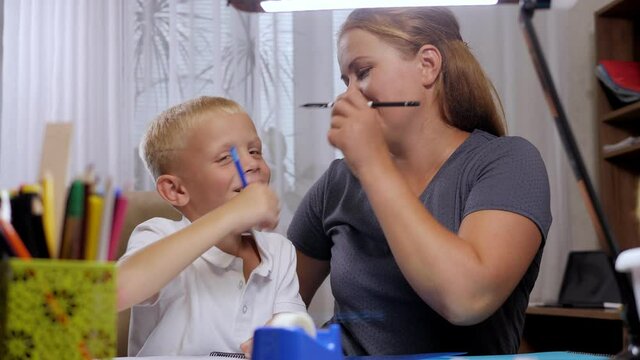 A happy mother and a little boy do homework together at home at the table, they have fun and paint each other's faces.