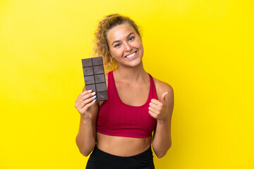 Girl with curly hair isolated on yellow background taking a chocolate tablet and with thumb up