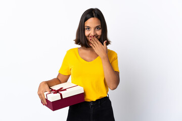 Young latin woman holding a gift isolated on white background happy and smiling covering mouth with hand