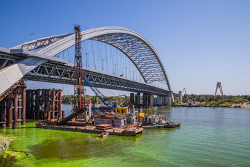 Fototapeta premium Construction of a large arched bridge in the capital of Ukraine. Podolsky bridge, panoramic view of the bridge under construction across the Dnipro, clear weather, summer. Kyiv