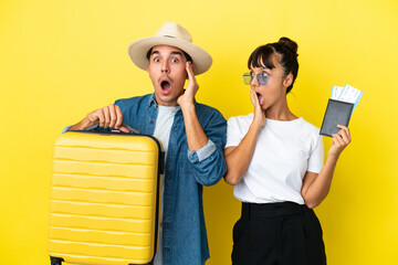 Young traveler friends holding a suitcase and passport isolated on yellow background with surprise and shocked facial expression