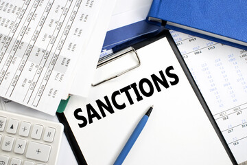 The word sanctions is written on a stationery tablet that lies on a financial document near a blue notebook. Political and economic concept