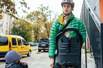 European father in protective helmet and green jacket taking his son after school or kindergarten at home with bicycle. Happy family concept.