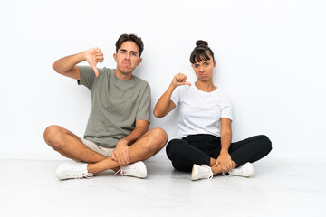 Young mixed race couple sitting on the floor isolated on white background showing thumb down sign with negative expression