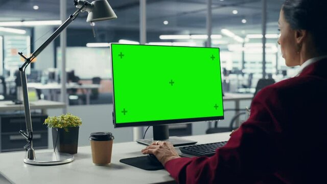 Successful Caucasian Businesswoman Sitting at Desk Working on Green Screen Laptop Computer in Office. Beautiful Businessperson using Chroma Key Display. Over Shoulder