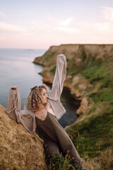 Curly woman posing at the edge of a rock above the sea, with blue sky and sea background. Autumn style. Travel and adventure.