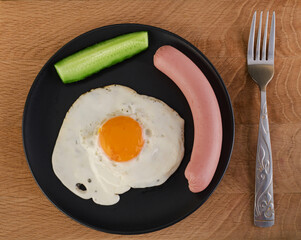 Scrambled eggs with chicken egg, sausage and a piece of cucumber on a black plate and cutlery on a wooden board