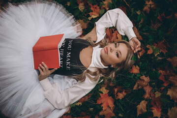 A girl in a white dress and a red book in her hands lies on colorful autumn leaves in the park