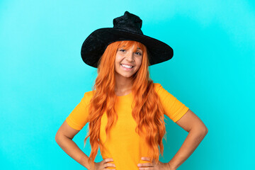 Young woman disguised as witch isolated on blue background posing with arms at hip and smiling