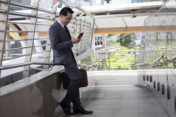 Asian businessman using mobile phone and smile outside of office in urban city. Asian man holding smartphone for business work.