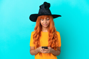 Young woman disguised as witch isolated on blue background sending a message with the mobile