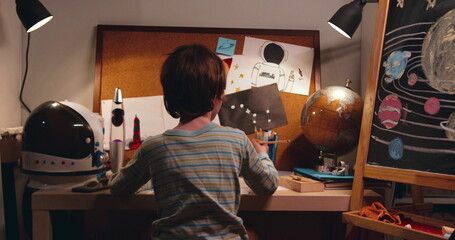 Cinematic rear view, little cute boy child takes pencil from glass to draw. Dreaming about future in space themed room.