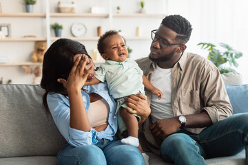 Tired black parents sitting with crying kid on sofa