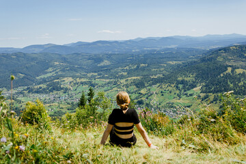 Little girl in a black T-shirt sitting on the green green meadow in Caprathian mountains. Back view. Travel, enjoying life, admire the beautiful nature view.
