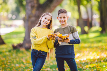 Happy twins teenagers boy and girl posing hugging each other in autumn park holding fallen yellow leaves in hand in sunny weather. Autumn season theme. Brother and sister have fun playing with leaves