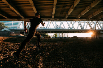 Man in a sports uniform under an overpass. Athlete runs forward under the overpass against the background of the sunset sky. There are bridges, roads and an industrial environment around the runner