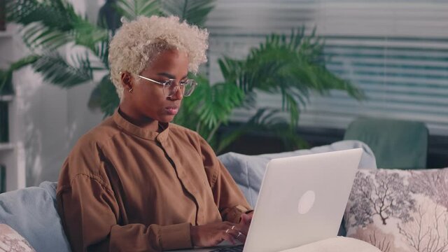 Concentrated African American woman sit on sofa busy working on laptop browsing web or checking mail. Focused bicultural young female typing texting on modern computer use wireless internet.