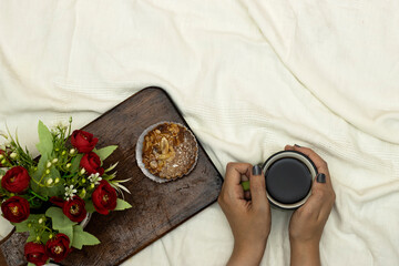 hands holding a cup of hot coffee with almonds pie, flowers pot on white bed background, vintage tone, top view. Lifestyle concept