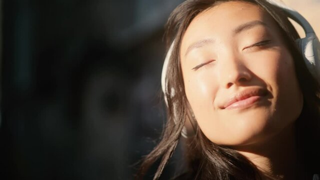 Close up of young millennial asian girl enjoying music in the city centre. Attractive young woman listening to music in headphones. High quality 4k footage