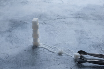 A pyramid of cubes of white sugar, a lane of granulated sugar from a crushed sugar cube, clamped with refined sugar tweezers. Selective focus.