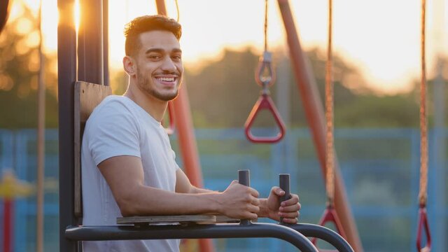 Portrait attractive millennial Middle Eastern Arab athletic guy doing exercise outdoor at sports ground. Young adult Indian sport man smiling affably standing beside equipment during morning workout