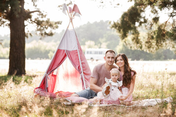 Mom, dad and little daughter are sitting next to wigwam decoration in the park. Family spending time outdoor in summer, having fun together. Girl are dressed in pink dress. © Andriy Medvediuk