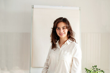 Portrait of young woman posing near blank empty whiteboard. Presentation concept. Teacher in white, businesswoman standing with a marker near a whiteboard, Online classes, business training 