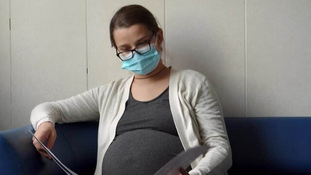 Young Pregnant Woman In Medicine Protective Mask Reading On Couch Waiting For Appointment Medical Control On Hospital. Visit Gynecologist Doctor At Clinic. Examine Expectant Belly Healthcare Check Up
