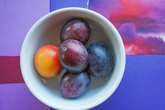 Purple plums and orange pluot from a fall farmers market, in white bowl on collage background with red and violet tones.