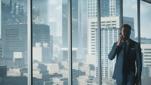 Confident Young Businessman in a Suit Walking in Modern Office, Talking on a Phone, Looking out of the Window on Big City with Skyscrapers. Successful Finance Manager Planning Work Projects.