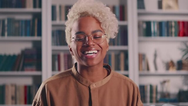Happy stylish African American gen millennial female student with blonde hair looks at camera stands indoors at home library. Smiling bicultural young woman, headshot portrait.