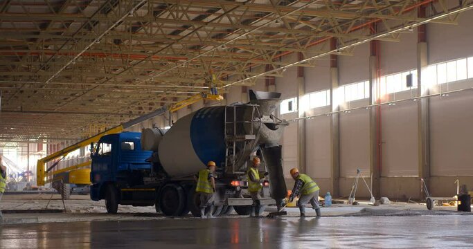 At a construction site in a workshop under construction, an auto concrete mixer delivers mortar for pouring the floor, workers level the concrete surface 