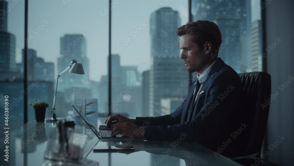 Poster confident businessman in a suit sitting at a desk in modern office, using laptop computer, next to w